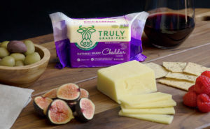 An image of a pack of Truly Grass Fed Cheddar Cheese and other pairing items like grapes and fruit
